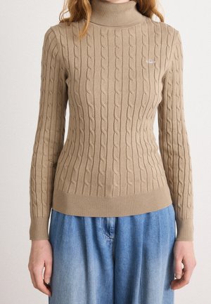 Pull à col roulé beige en maille torsadée porté avec un jean bleu ample sur une personne debout.