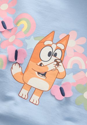 Un tessuto azzurro chiaro presenta un'illustrazione di un cane arancione in stile cartone animato con grandi occhi, circondato da fiori colorati, arcobaleni e farfalle tridimensionali.