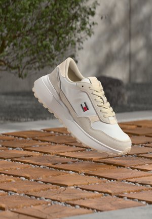 Beige and white sneaker with red, white, and black logo, mid-air above wooden tile floor with pebbles and blurred outdoor background.
