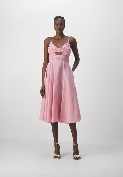 Roze jurk met een aansluitend lijfje met een strik en uitsparing, een uitlopende rok en dunne bandjes. Gemaakt met witte schoenen met hoge hakken.