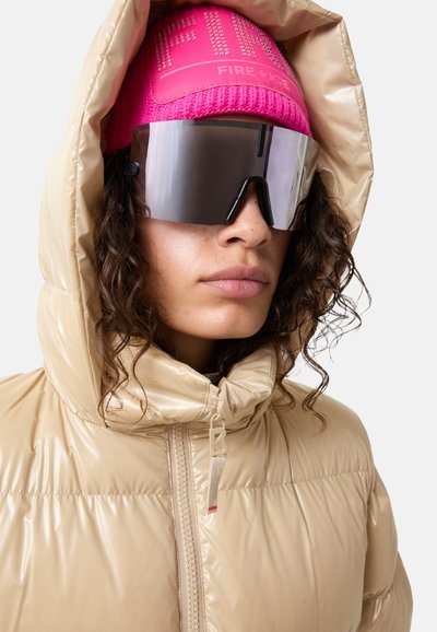Beige pufferjakke med en blank overflade og snoretræk i huen, parret med en strålende pink strikbeanie og store sorte solbriller.