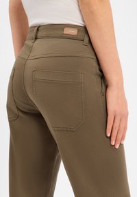 Pantalon vert olive avec une texture lisse, poches arrière, et une étiquette en cuir sur la ceinture. Le design présente une coupe ajustée.