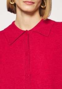 Femme portant un pull boutonné rouge vif avec un large col et de grandes boucles d’oreilles en or, sur un fond clair uni.