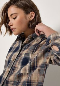 Flannel shirt with a blue, beige, and brown plaid pattern, featuring a button-down collar, chest pocket, and wooden buttons.