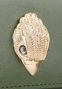 Fermaglio metallico dorato a forma di foglie sovrapposte con un dettaglio in pietra nera sfaccettata, su uno sfondo verde oliva texturizzato.