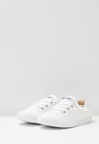 White canvas sneakers with textured upper, lace-up design, rubber soles, and minimalistic branding on tongue. Smooth interior lining.