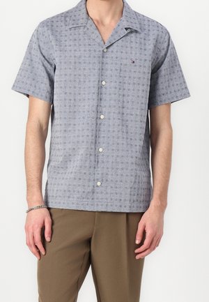 Mann trägt ein hellblaues, kurzärmeliges Hemd mit feinem geometrischem Muster und braune Hose, steht vor weißem Hintergrund.