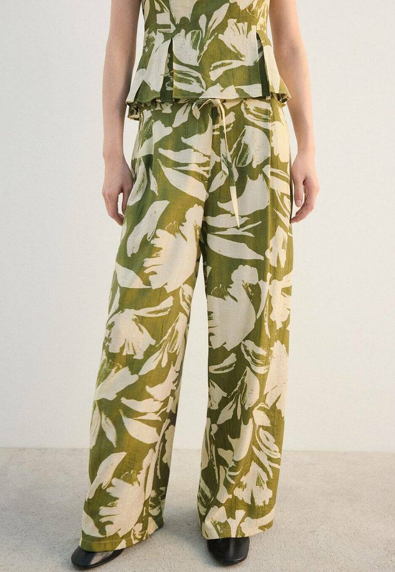 Pantalon à jambes larges à motif floral vert et crème avec taille à cordon, en tissu léger. Des chaussures noires complètent la tenue.