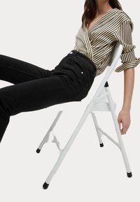 Bílá skládací židle s moderním designem, která má pevný rám a černé gumové nožičky. Modelka má na sobě pruhované tričko a černé džíny.