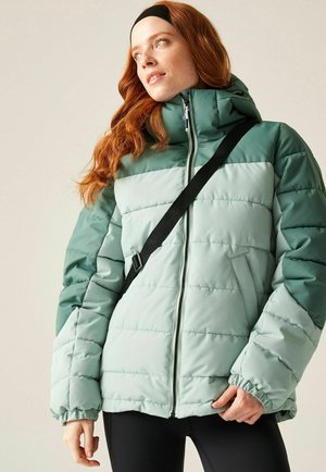 Kobieta o rudych włosach, ubrana w zieloną i jasno-miętową pikowaną kurtkę, czarną opaskę na głowę oraz czarny pasek na ramię, spogląda na bok.