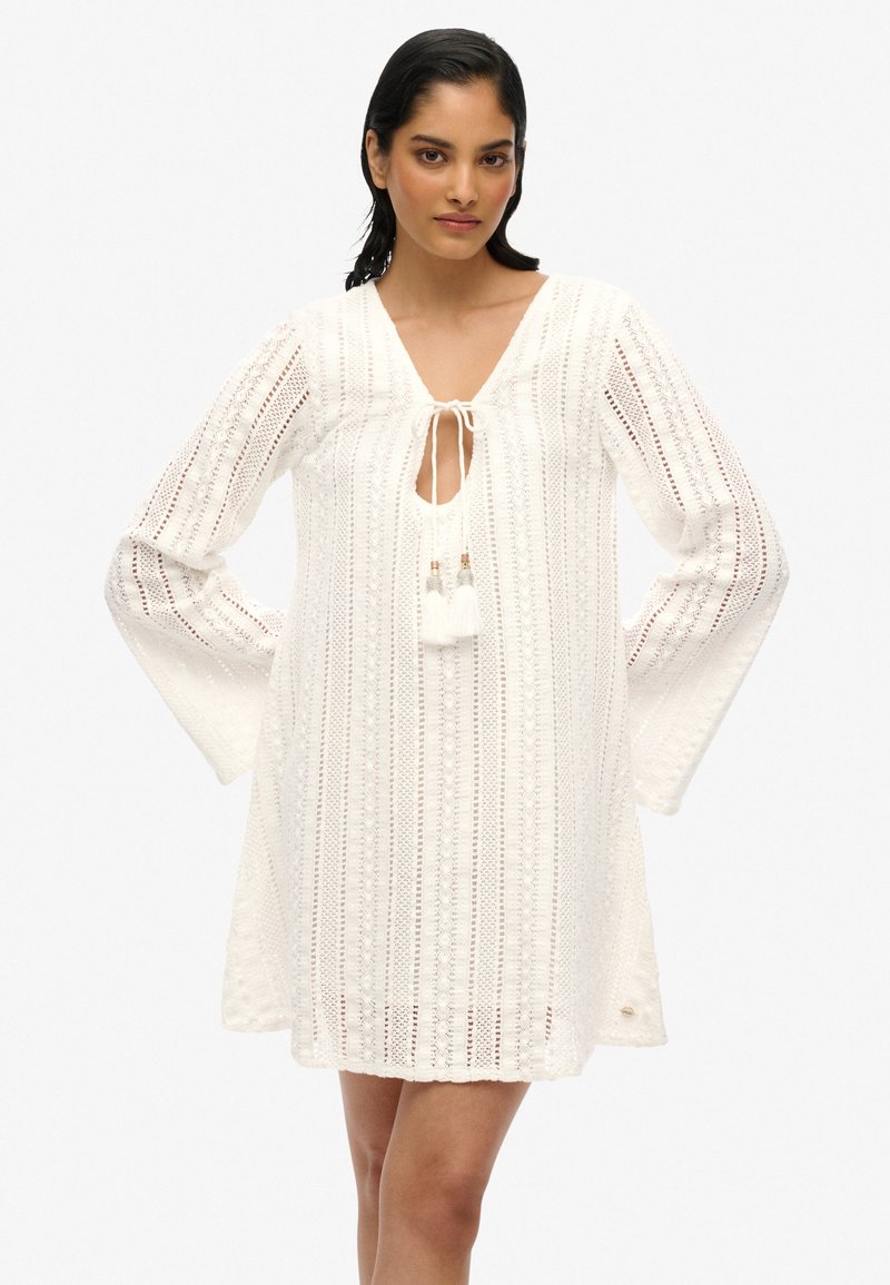 Vestido blanco hasta la rodilla con escote en V, mangas largas y un lazo en el escote, con patrones texturizados y acentos de borlas.