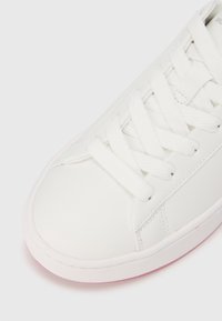 Λευκό αθλητικό παπούτσι με λείο δερμάτινο επάνω μέρος, στρογγυλεμένη μύτη και χοντρές λευκές κορδόνια. Διαθέτει υφή στη σόλα με μια ελαφριά ροζ απόχρωση στο κάτω μέρος.