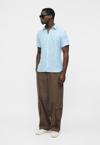 Man wearing sunglasses, light blue short-sleeve striped shirt, loose brown cargo pants, and white sneakers, standing against a white background.