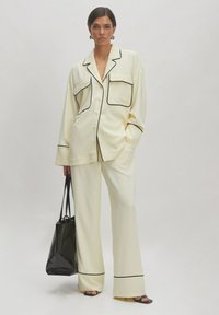 Ensemble de pyjama deux pièces crème avec piping noir, comprenant une chemise ample avec poches et un pantalon large, accompagné d'un sac fourre-tout noir.