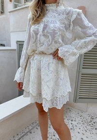 Blouse en dentelle blanche avec un col et des manches à volants, associée avec une mini-jupe en dentelle à couches, toutes deux ornées de motifs floraux et de bords festonnés.