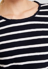 Maglietta a righe blu navy e bianca con scollatura rotonda. Il tessuto sembra morbido con una leggera texture e bordi leggermente sfrangiati.