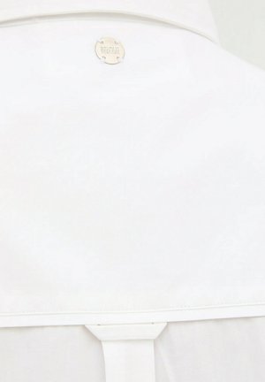 Tessuto bianco con bordo cucito e un emblema metallico rotondo etichettato "SAFARI BEACH" vicino al centro superiore.