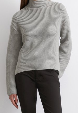 Žena nosící světle šedý žebrovaný pletený rolák a černé kalhoty, stojící před jednobarevným bílým pozadím.