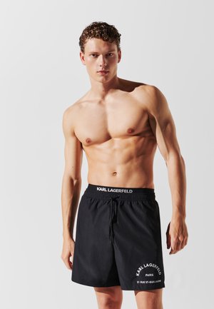 Pantaloni scurți de baie negri cu un elastic la talie, având accente albe de logo și text. Textură ușoară și netedă, design până la genunchi.