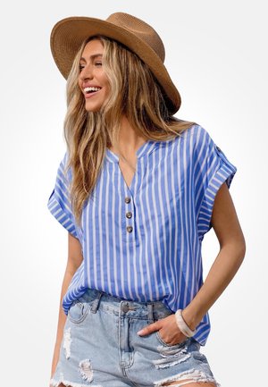 Lachende vrouw met lang blond haar, een brede bruine hoed, blauw-wit gestreepte blouse met korte mouwen, gescheurde spijkershorts en een armband.