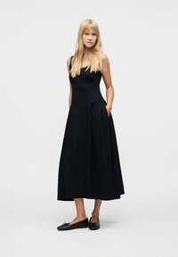 Γυναίκα με μακριά ξανθά μαλλιά, που φοράει μαύρο μίντι φόρεμα χωρίς μανίκια με τσέπες και μαύρα μπαλαρίνια, στέκεται μπροστά σε λευκό φόντο.