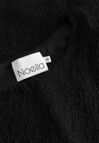 Čierna textúrovaná látka šiat s zložitým vzorom, obsahujúca hladkú čiernu podšívku a štítok s nápisom "Noella S."