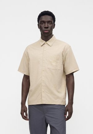 Homme portant une chemise beige à manches courtes avec une poche poitrine et un pantalon gris foncé, debout sur un fond blanc uni.