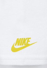 Žlté logo Nike so znakmi na bielom textilnom pozadí, umiestnené v strede blízko dolného okraja.