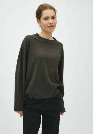 Jeune femme aux cheveux bouclés portant un pull à manches longues vert foncé et un pantalon noir, debout devant un fond clair uni.