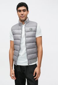 Siva puhasta jopica z navpičnimi prešivi, sprednja zadrga in značko z logotipom. Nosi se preko svetlo sive polo majice, združena s temnimi hlačami.