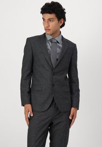 Costume gris à motifs avec une veste ajustée, deux boutons et des revers en pointe. Associé à une chemise habillée grise et une cravate. Texture de tissu lisse.