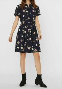 Blumenkleid aus marineblauem Stoff mit kurzen Puffärmeln, durchgehender Knopfleiste und elastischem Taillenbund. Kombiniert mit schwarzen Stiefeletten.