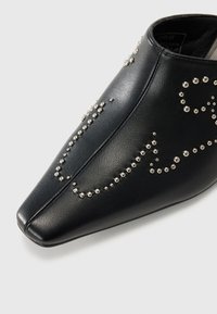 Chaussure pointue en cuir noir avec des clous argentés décoratifs disposés en motifs tourbillonnants sur la surface supérieure.