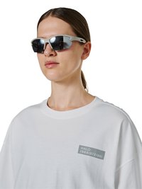 Junge Person mit braunen, zurückgebundenen Haaren, trägt weißes T-Shirt mit kleinem Text und graue Sportsonnenbrille mit verspiegelten Gläsern.