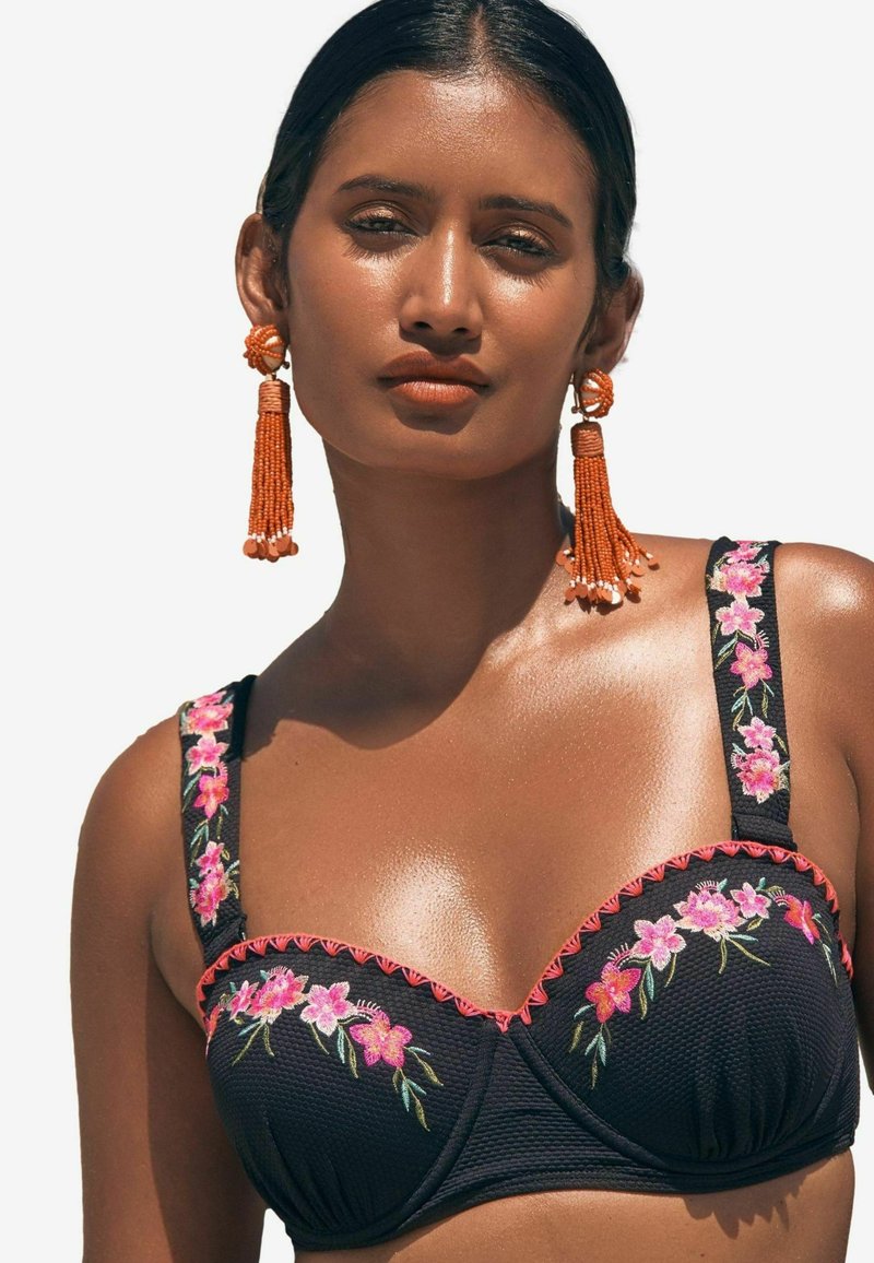 Reggiseno nero con ricami floreali in rosa e verde, caratterizzato da spalline larghe con bordi ondulati. Modella che indossa orecchini di frange arancioni.