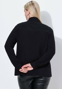 Sort langærmet bluse med en knappet manchet og en glat tekstur. Har et kontrastpanel ved udskæringen og en lige kant.