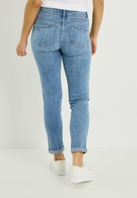 Vaqueros slim fit azul claro con bolsillos traseros, detalles bordados y puños remangados, combinados con zapatillas blancas sobre un fondo neutro.