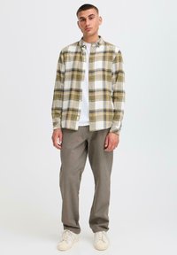 Geruit flanellen overhemd in olijfgroen, beige en wit, met een knoopsluiting. Gecombineerd met grijze broek en witte sneakers.