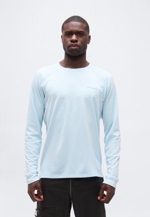 Young man standing facing forward wearing a light blue long-sleeve Columbia shirt and black pants against a plain background.