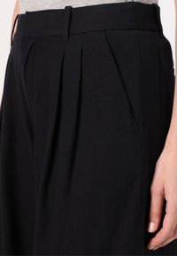 Pantalons plissés noirs en tissu léger, avec une taille ajustée et des poches latérales ; texture lisse avec un design structuré.