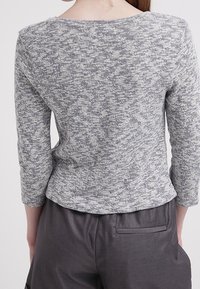 Grau gestrickter Oberteil mit langen Ärmeln, Rundhalsausschnitt und strukturiertem Muster. Trägt eine graue Hose mit einer Gesäßtasche.