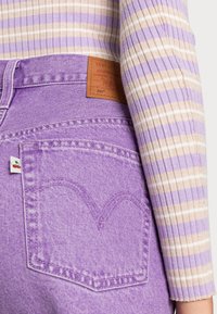 Vaqueros de denim morado con un detalle de costura en forma de corazón en el bolsillo trasero y una etiqueta de marca de cuero marrón. Top a rayas en lavanda y beige.