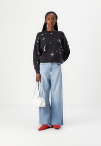 Zwarte trui met hemelse patronen, gecombineerd met wijde lichtblauwe jeans. Een witte handtas en rode schoenen maken de outfit af.