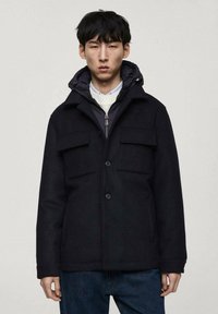 Svart ullblandad kappa med dragkedja vid kragen, tryckknappar och fyra framfickor. Har en lager-på-lager huva och en avslappnad passform.