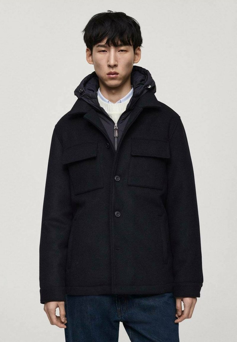 Svart ullblandad kappa med dragkedja vid kragen, tryckknappar och fyra framfickor. Har en lager-på-lager huva och en avslappnad passform.