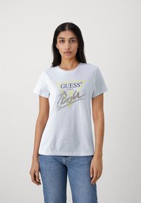 Camiseta blanca de algodón con un gráfico de triángulo amarillo y "GUESS" en la parte superior, con una firma adornada con strass en el centro.