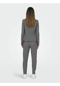 Grå skräddarsydd kostym med en figurnära jacka och matchande veckade byxor. Personen bär vita sneakers. Slät tygtextur, minimalistisk design.