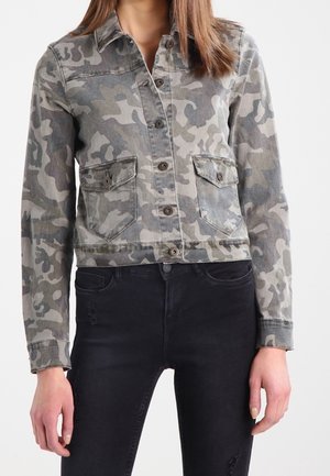 Kvinde iført en kort grå camouflagejakke med knapper og to forlommer samt sorte tætsiddende jeans.