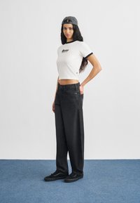 Γυναίκα που στέκεται με μακριά σκούρα μαλλιά, φοράει λευκό κοντομάνικο μπλουζάκι, μαύρο τζιν παντελόνι με φαρδιές ποδές, μαύρα αθλητικά παπούτσια και γκρι καπέλο φοράει ανάποδα.