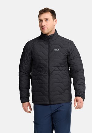 Mann trägt eine schwarze gesteppte Jacke mit Reißverschluss und hohem Kragen sowie blaue Hose, steht vor einem einfarbigen hellen Hintergrund.